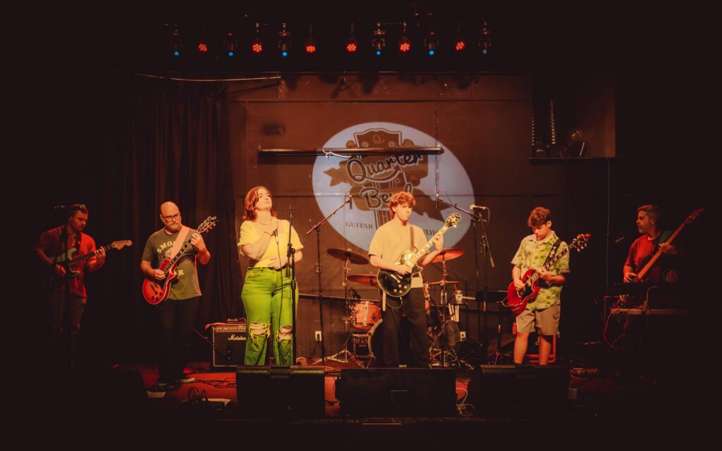 Six Quarter Bend guitar students performing on stage under the studio logo at a Mega Concert in Lancaster, Pennsylvania